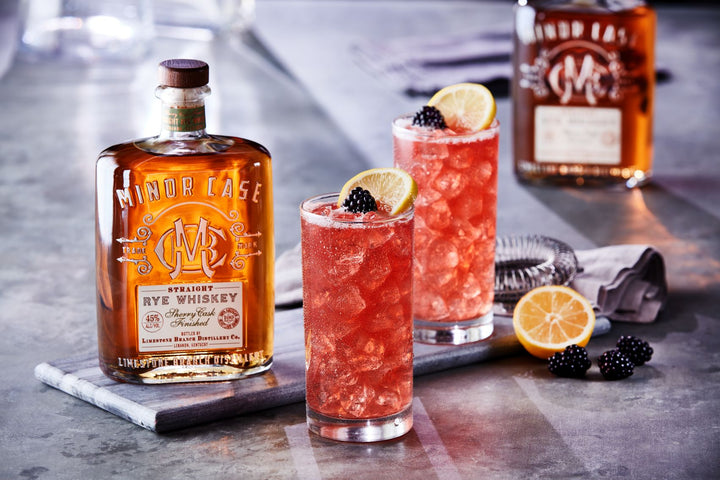 Two cocktails with lemon slices and blackberries next to a bottle of Minor Case whiskey on a bar.