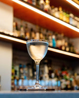 A Mizu Martini shown in an elegant, iconic setting. Clear sumptuous liquid in a NIck & Nora Glass. The background is a large tall shelf of premium spirits, blurred and out of focus. The light is warm and inviting.