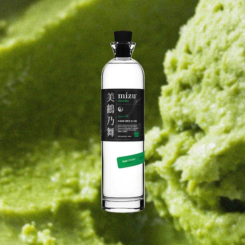 A lovely tall thin bottle with a black cap. The label is black with Japanese characters on the left side. It reads in English: Mizu Shochu Green Tea. This is against a macro shot of delicious looking matcha sorbet. 