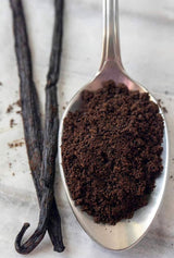 Silver spoon filled with ground coffee next to two vanilla beans on a light background