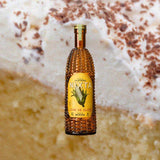 A bottle of rich yellow looking alcohol. The bottle itself has the shape and texture of a corn cob. The label reads: Nixta Licor de Elote. Hecho En Mexico.  This is set against a backdrop of a tres leches cake, with a thick layer of whipped cream frosting on top.