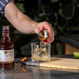 Person pouring a drink from a bottle into a glass with a lime and wooden cutting board on a bar.