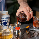 Person pouring a liquid from a bottle into a metal jigger on a bar counter.