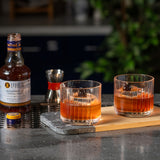 Two glasses of amber-colored cocktails on a bar with a bottle of Liber & Co Old Fashioned Cocktail Syrup.