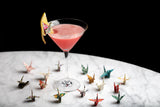Pink cocktail in a martini glass with lemon twist on a marble surface, surrounded by colorful origami butterflies.