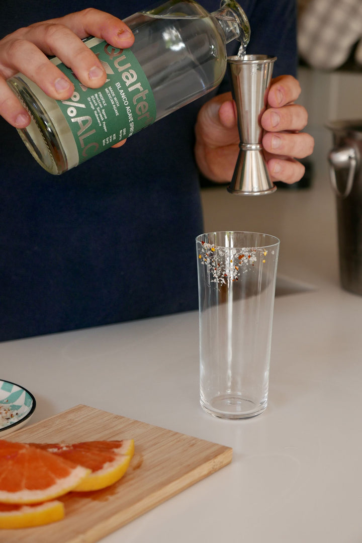 Person pouring a drink from a bottle into a glass with a jigger, surrounded by orange slices on a cutting board.