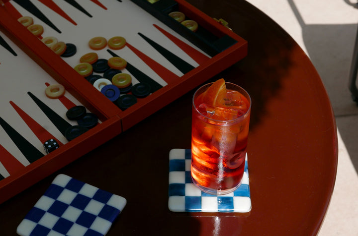 Glass of red cocktail with orange slice on a checkered coaster next to a backgammon board.