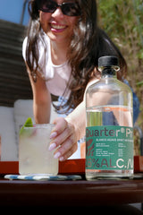 Woman holding a drink with a bottle of Quarter Proof Agave Spirit on a table outdoors.