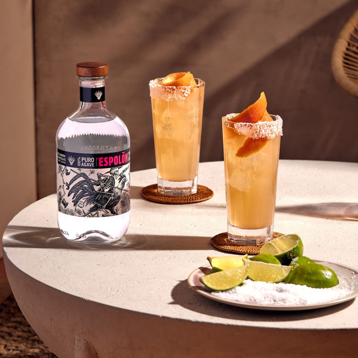 Two cocktails with orange slices and limes on a plate, next to a bottle of Espolón tequila.