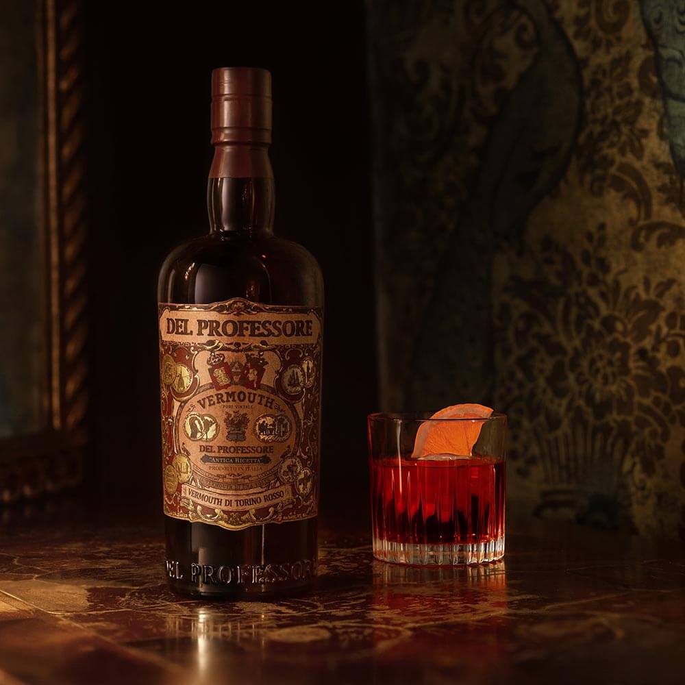 A textured sumptuous background of gilded peacock wallpaper. The stately bottle of Del Professore Vermouth di Torino Rosso sits in the foreground, a delicious looking negroni right next to it.