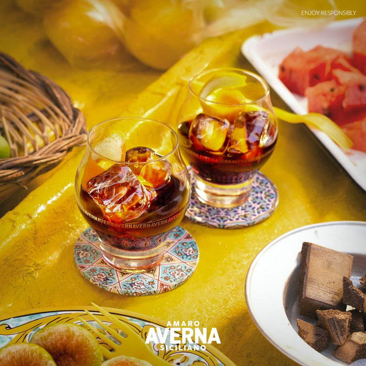 Two glasses of Amaro Averna with ice cubes on a yellow tablecloth, surrounded by fruits and cookies.