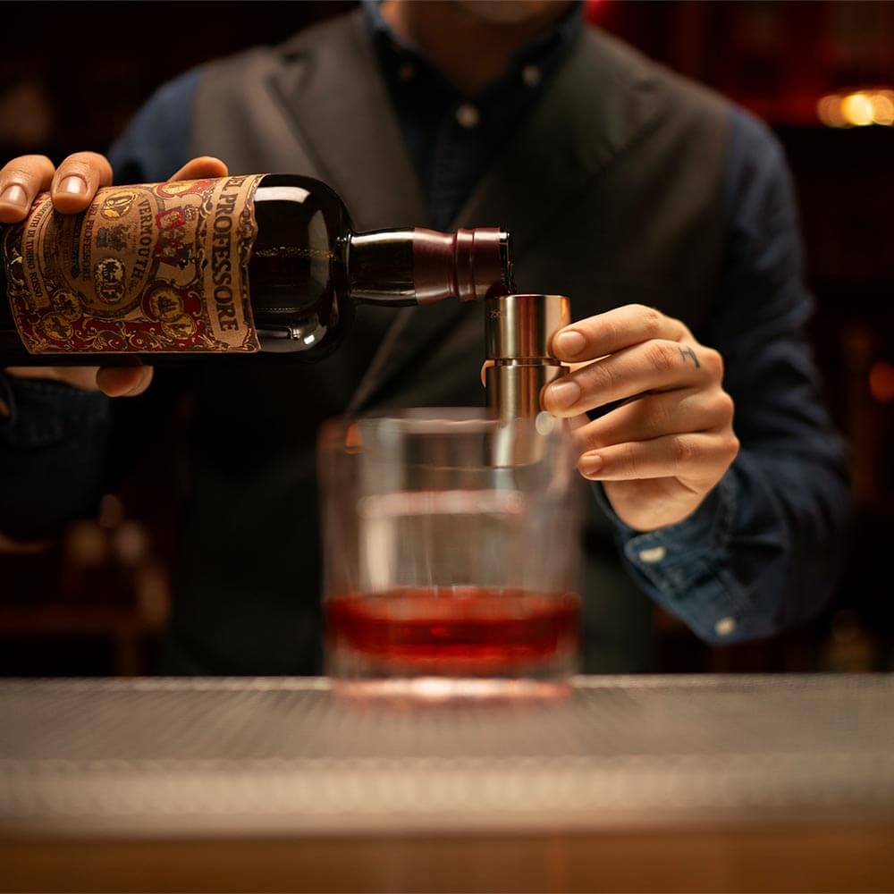 A close-up shot of a bartender pouring from a bottle of Del Professore Vermouth di Torino Rosso. There's a Negroni glass in the blurred foreground.