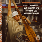 An image of a man stirring a giant vat of roasted chiles in their own juices with a large wooden paddle.  The words, "Crafted in Puebla and inspired by a 100-year-old Menjurje recipe." are in the upper right corner.