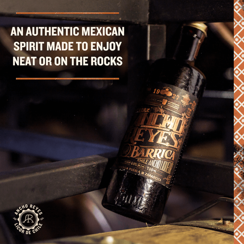 A bottle of Ancho Reyes Barrica tipped on its side against a steel bar.  "An authentic Mexican Spirit made to enjoy neat or on the rocks" in the upper left corner.