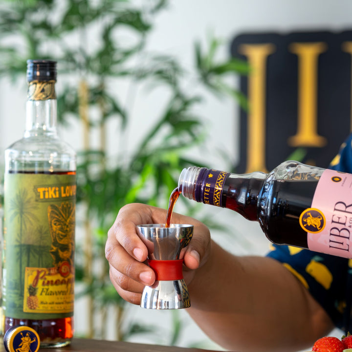 Person pouring liquor from a bottle into a cocktail shaker with bottles of Tiki Love and Liber on a table.