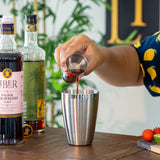 Person pouring liquid from a measuring cup into a cocktail shaker with bottles and ingredients in the background.