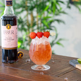 Cocktail with strawberries and a bottle of Liber Pacific Strawberry Syrup on a wooden surface.