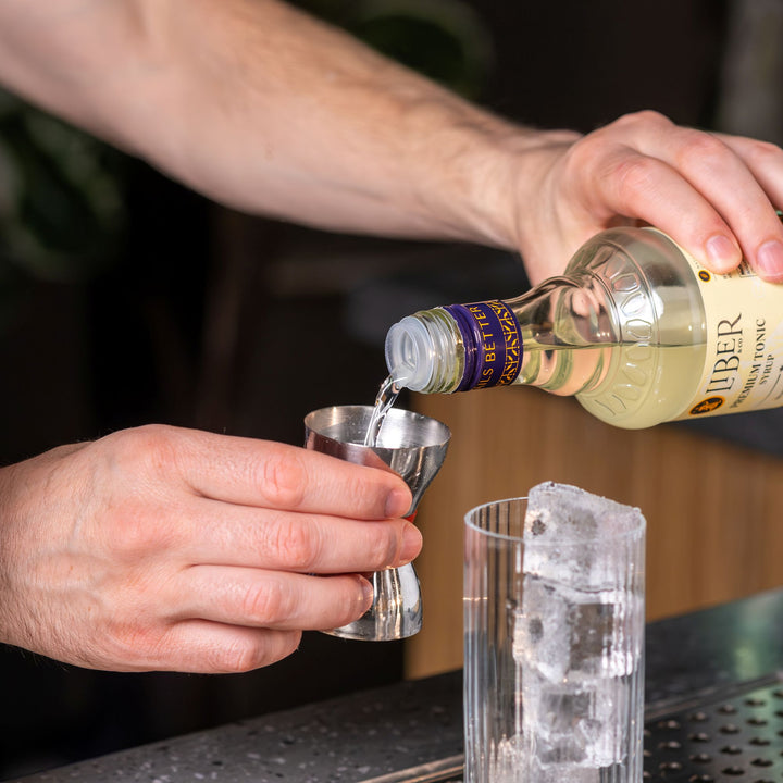 Person pouring a clear liquid from a bottle into a metal jigger with ice cubes in the background.