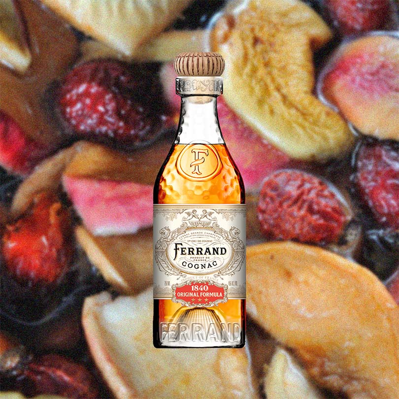 A bottle of Pierre Ferrand Original Formula with a clear amber-colored liquid visible and the brand's logo on the label over backdrop of fruits.
