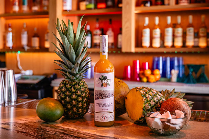 Bottle of Chinola pineapple liqueur on a bar counter with a pineapple and other fruits.