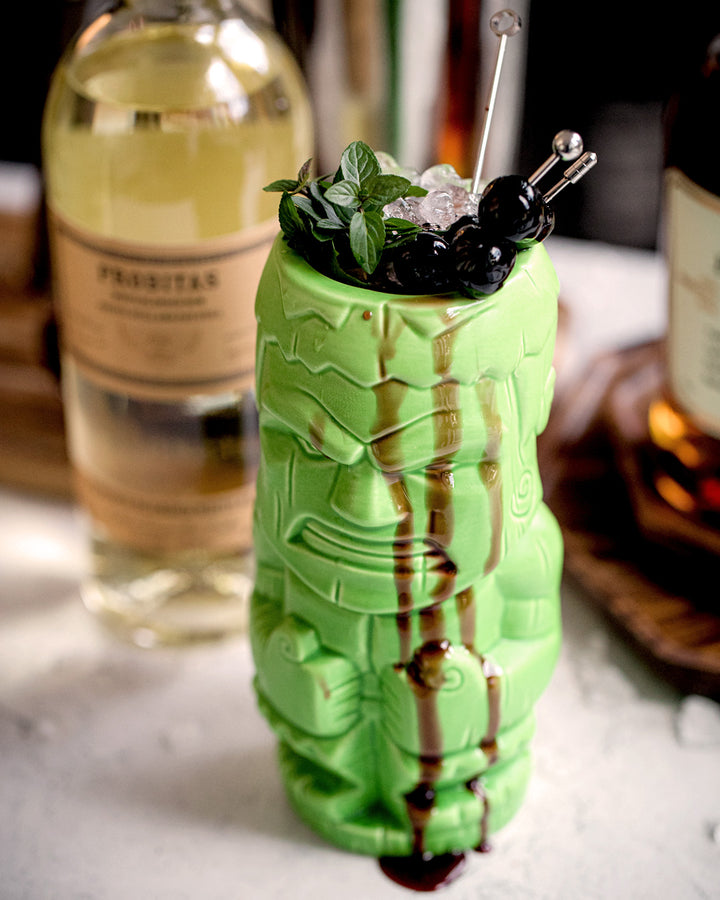 Green tiki mug with a cocktail on a bar counter, featuring a bottle of Prosecco in the background.