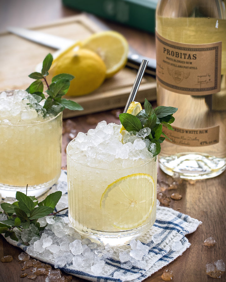 Two glasses of lemonade with ice and garnishes on a wooden table with a bottle of Prohibitas in the background.