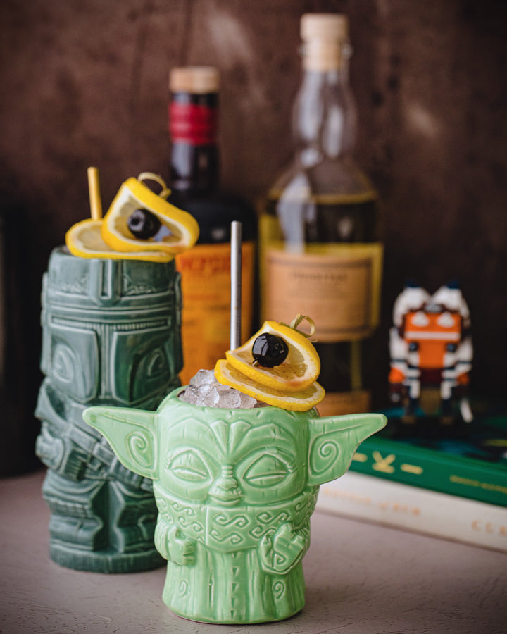 Two green tiki mugs with drinks and bottles in the background