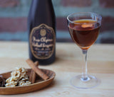 Bottle of Nux Alpina Walnut Liqueur with a glass of red wine, nuts, and cinnamon sticks on a wooden surface.