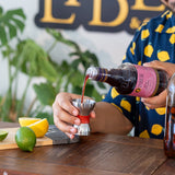 Person pouring a dark liquid from a bottle into a silver cocktail shaker on a wooden table with lemons and limes.