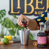 Person pouring a red liquid into a cocktail shaker with a 'Libe & Co' sign in the background.