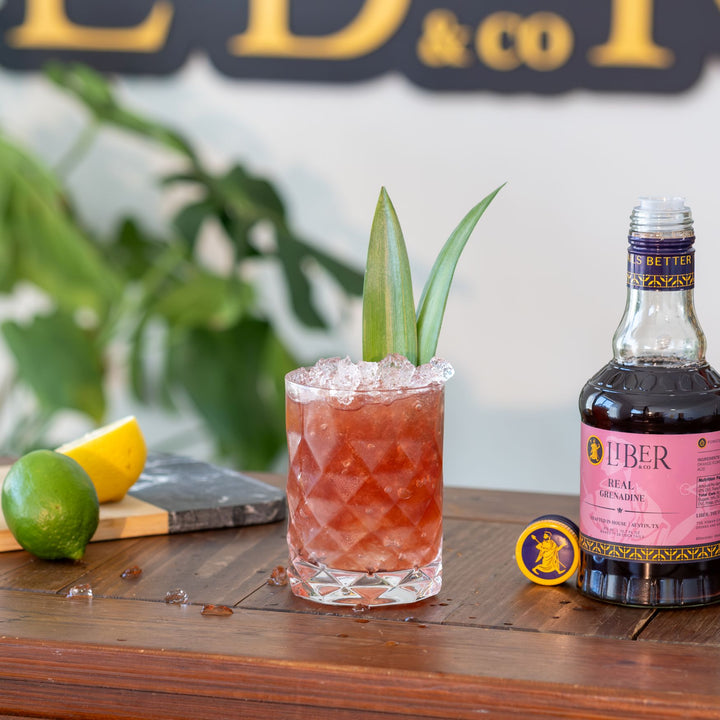 Cocktail with ice and garnish next to a bottle of Liber & Co Real Grenadine on a wooden surface.