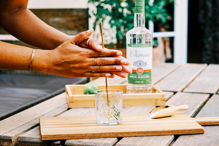Person preparing a drink with a bottle of Clement Blanc on a wooden table outdoors.