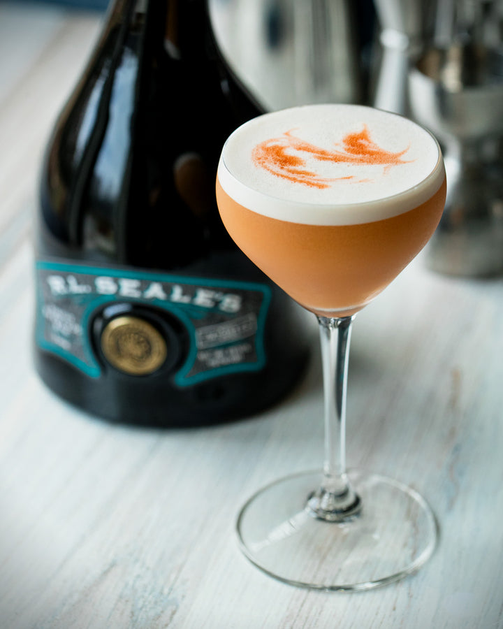 Cocktail in a glass with a bottle of El Seale's whiskey in the background on a wooden surface.