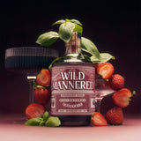 A bottle of Wild Mannered Strawberry Basil All-Natural Cocktail surrounded by fresh strawberries, basil leaves, and two dark stemmed glasses on a dark pink background.