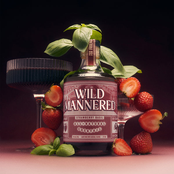 A bottle of Wild Mannered Strawberry Basil All-Natural Cocktail surrounded by fresh strawberries, basil leaves, and two dark stemmed glasses on a dark pink background.