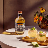 Espolón tequila bottle with two cocktails and a vase of flowers on a table.