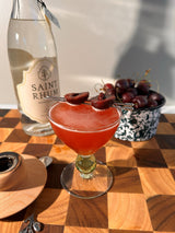 Cocktail with cherries in a glass on a wooden table next to a bottle of Saint Rhum.