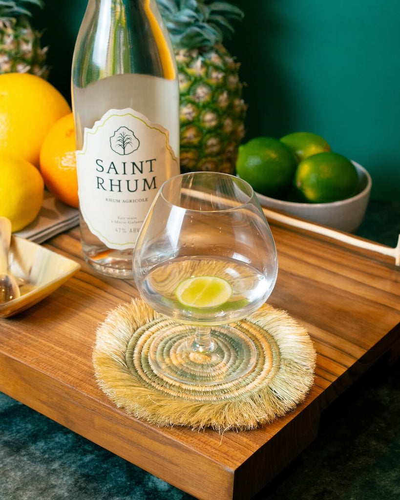 Bottle of Saint Rhum Rum on a wooden tray next to a glass of rum with a lime garnish with fruit in the background.