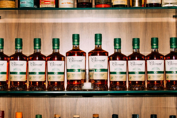 Row of Clement brand bottles on a wooden shelf with various other bottles in the background.