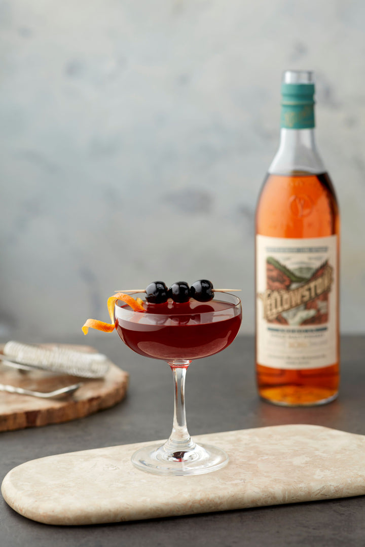 Cocktail in a glass with garnishes next to a bottle of Wild Turkey bourbon on a stone surface.