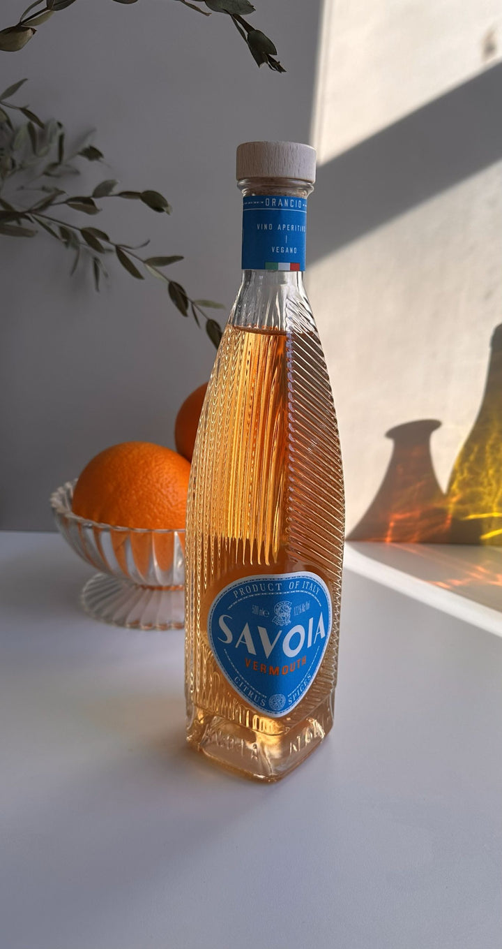 Bottle of Savoia brand on a white surface with an orange and decorative items in the background.