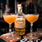 Two cocktails in glasses with a bottle of Sihenge Pineapple Rum on a bar counter.