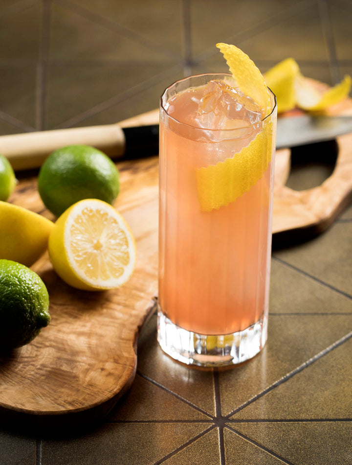 Cocktail with lemon and lime garnish on a wooden cutting board.