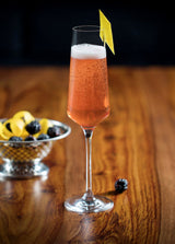Cocktail in a glass with a lemon twist on a wooden table, accompanied by a bowl of fruit.