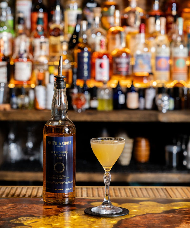 Bottle of Smith & Cross Rum and a cocktail glass with a drink on a bar counter, with shelves of bottles in the background.