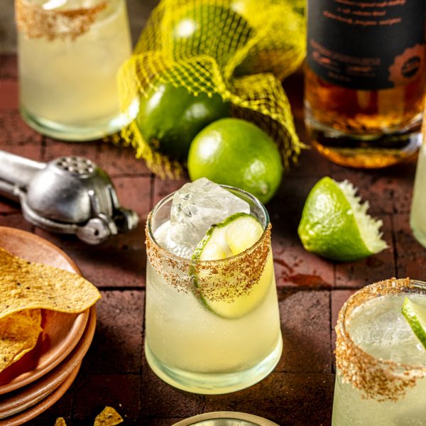 Cocktail with lime and ice in a glass on a wooden surface with limes and a bottle in the background.