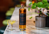 Bottle of whiskey on a marble surface with a cocktail and plant in the background