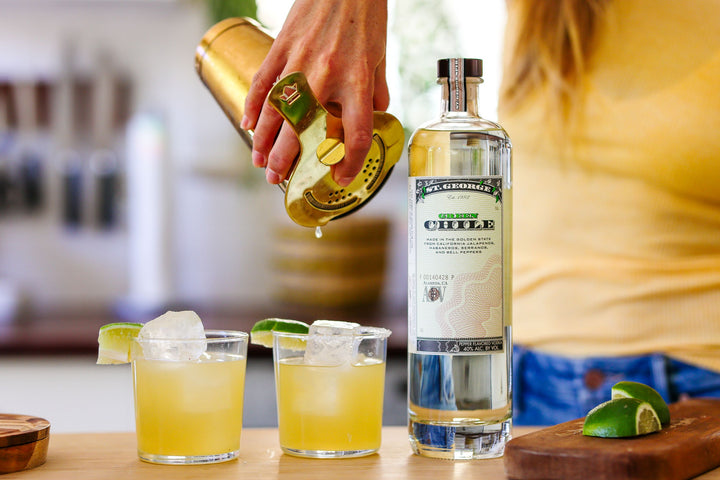 Person pouring a drink from a bottle labeled 'St. Germain' into two glasses with lime wedges on a wooden surface.