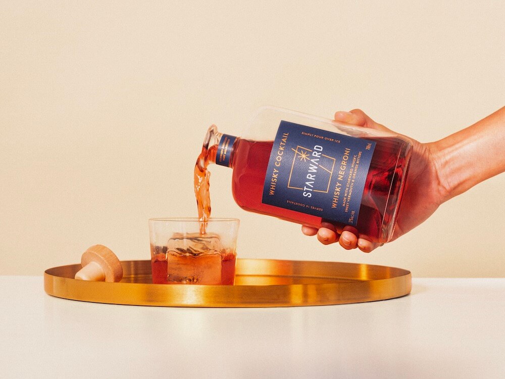 Someone pouring Starward Whisky Negroni into a glass featuring one large ice cube resting on a gold tray.