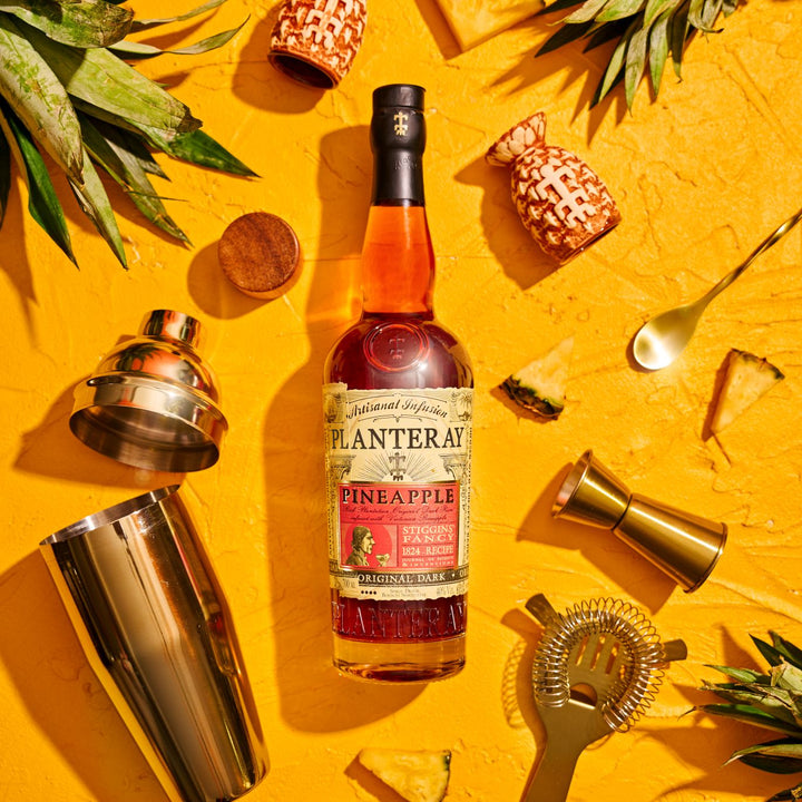 Bottle of Planteray Pineapple Rum on a yellow background with cocktail-making tools.
